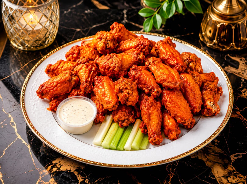 A large platter of buffalo wings with ranch dressing and celery sticks on a marble table.