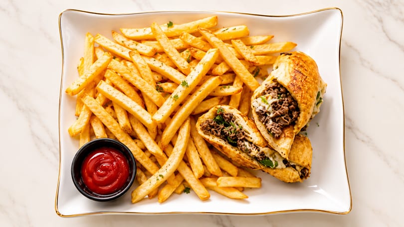 A white rectangular plate with golden fries, two cheesesteak halves, and a side of ketchup.